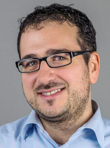 Portrait Giorgio Sangiovanni mit Brille und leichtem Lächeln vor grauem Hintergrund.